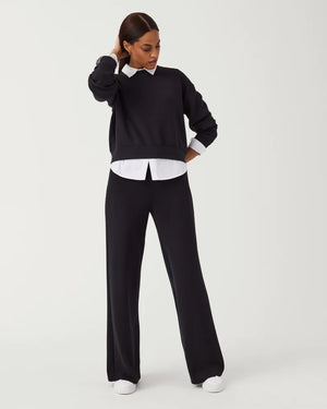 A woman stands against a plain white background, modeling the SPANX - AIRESSENTIALS WIDE LEG PANT in dark blue, paired with a long-sleeved, half-zip pullover made from lightweight fabric. She has her right hand touching the zipper near her chin and is wearing white sneakers. The relaxed fit adds to the ensemble's casual appeal.