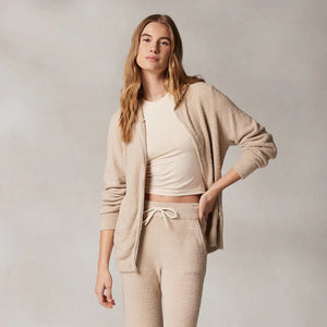 A person with long hair, wearing Kashwere's KASHWERE - RELAXED HOODIE made from Kash-ease yarn, has their hands in the front pockets of a light beige zip-up. The hood is up against a neutral, textured background. This wardrobe staple from KASHWERE looks both comfortable and casual.