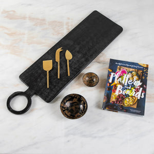 A black cutting board with a round handle displays three gold cheese knives of different shapes. Next to it are two Medium Horn Bowls by Be Home, Inc., speckled in brown, and a hardcover book titled "Platters and Boards" showcasing a colorful food spread on the cover—a perfect serving kit that's ethically crafted.