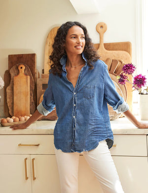 A person with curly hair stands in a kitchen, leaning back against a counter with hands resting on the surface. They wear an Eileen Relaxed Button Up Vintage Indigo Denim shirt from FRANK & EILEEN with rolled-up sleeves and white pants. The counter has a collection of wooden cutting boards and a vase with purple flowers.