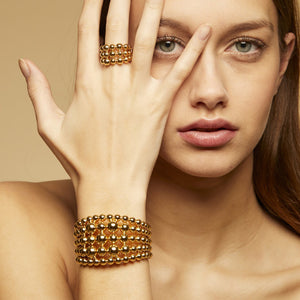 A person with long hair is partially covering their face with their hand, displaying a GAS BIJOUX - MUTIPERLA RING on their finger. They are also wearing a matching gold beaded bracelet on their wrist from the brand GAS BIJOUX. The background is a neutral beige color.