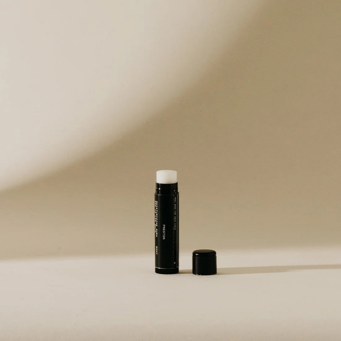 A black cylindrical tube of PRESTON - SUNSCREEN LIP BALM SPF15 stands upright on a beige surface with a shadow cast behind it. The cap is placed beside it on the right, and the hydrating beeswax balm is partially extended from the tube. The background is light beige and out of focus.