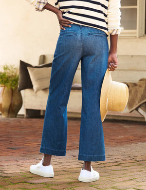 A person wearing a striped sweater, lightweight FRANK AND EILEEN - WESTPORT ITALIAN WIDE LEG TROUSER, and white sneakers stands on a brick path, holding a straw hat. The casual scene is enhanced by the textured backdrop, offering a relaxed yet stylish vibe.