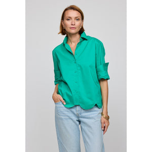 A person models the HINSON WU - BLAIRE SCALLOP HEM SHIRT in bright green with rolled sleeves, paired with light blue jeans, one hand in their pocket against a white background, highlighting the versatile styling of stretch cotton.