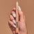 Close-up of a hand with white painted nails holding a sparkle pearl colored eyeshadow pencil with a pink tip, branded with "JASON WU BEAUTY" in black text against a solid neutral beige background.