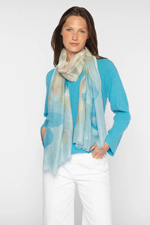 A person with long, dark hair stands against a plain background, elegantly wearing a KINROSS light blue cashmere sweater, white pants, and the KINROSS CASHMERE - KIWI BLOOMS PRINT SCARF.