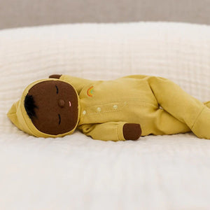 An adorable DOZY DINKUM DOLL by OLLI ELLA USA, featuring soft brown fabric skin, closed embroidered eyes, and short black hair, is dressed in a charming yellow onesie adorned with a rainbow design on the chest. Perfect as a first toy, the doll is lying on a white, quilted surface.