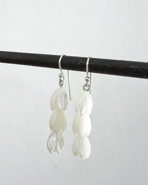 A pair of ANN LIGHTFOOT - TRIPLE TULIP MOTHER OF PEARL EARRINGS hangs from a black rod against a plain light gray background. Each earring features three petal-shaped segments stacked vertically, creating a cascading effect, and is adorned with sterling silver earwires for added elegance.