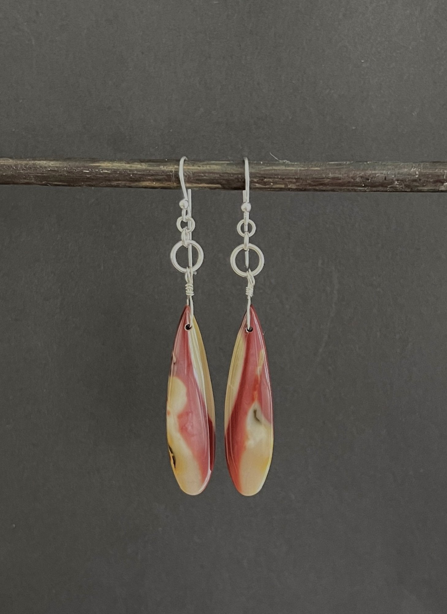 The ANN LIGHTFOOT Mookite Drop Earrings boast a long, teardrop shape with a stunning marbled design in vibrant red and white hues. Mounted on sterling silver hooks, they are beautifully showcased on a pristine white background.