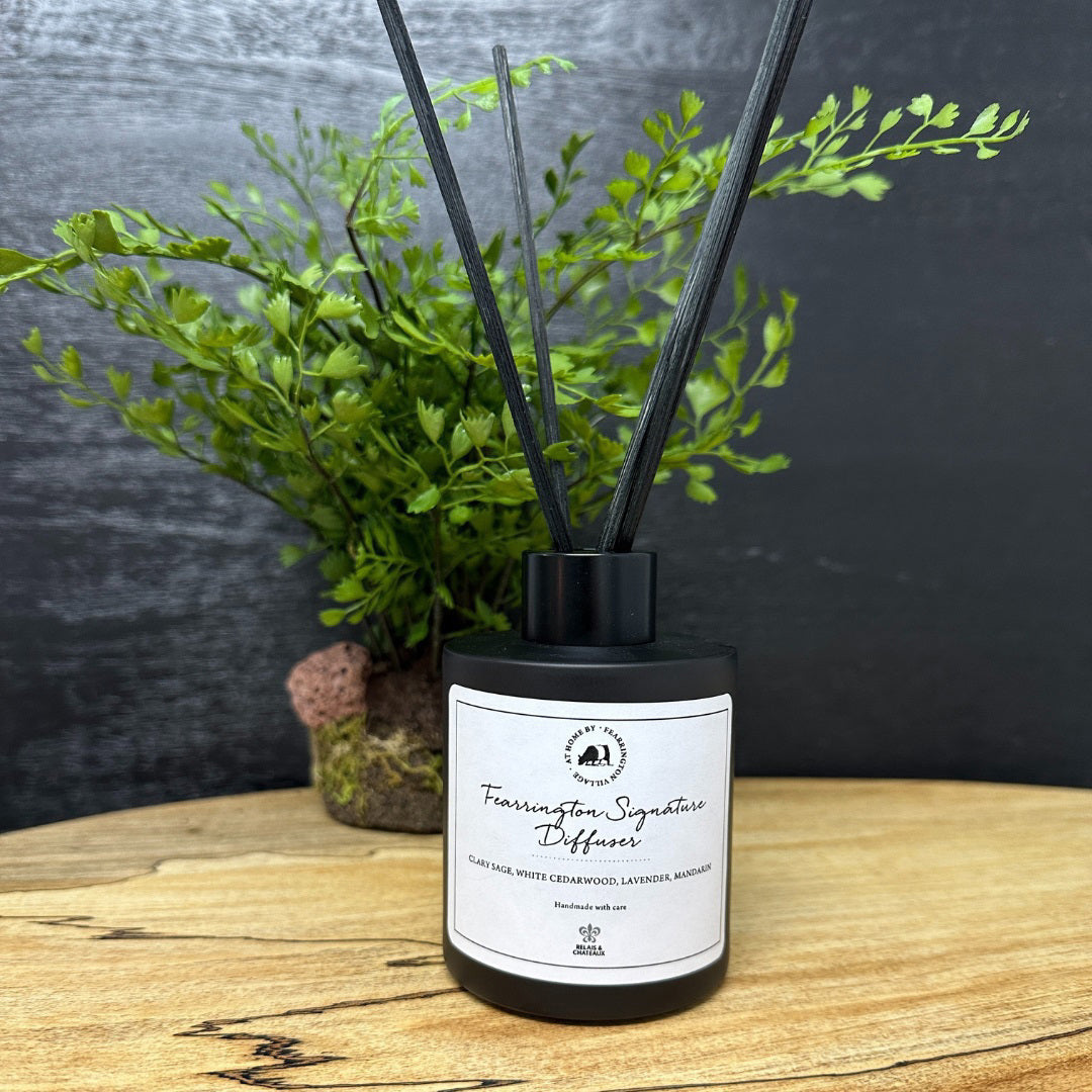 A matte black glass bottle containing several thin black reeds makes an ideal holiday gift. The label on the front reads "Fearrington Signature Diffuser - Clary Sage, White Cedarwood, Calming Lavender, Marjoram." The backdrop is a white, textured surface.