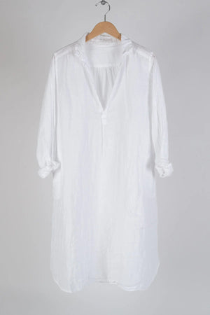 The CP SHADES - LARA LINEN DRESS in white, with long sleeves, hangs casually on a wooden hanger against a plain gray background. Made from 100% linen, this dress shirt features a deep V-neckline with a single button closure and possesses a relaxed, slightly wrinkled fabric. The sleeves are nonchalantly rolled up to the elbows, reflecting an essence of eco-friendly fashion synonymous with the CP SHADES brand.