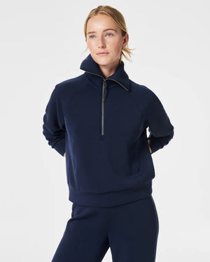 A person with short hair is wearing a SPANX AIRESSENTIALS HALF ZIP sweater in navy blue, made from lightweight spacer fabric, paired with black pants. Their arms are crossed over their chest as they stand against a plain, light grey background.