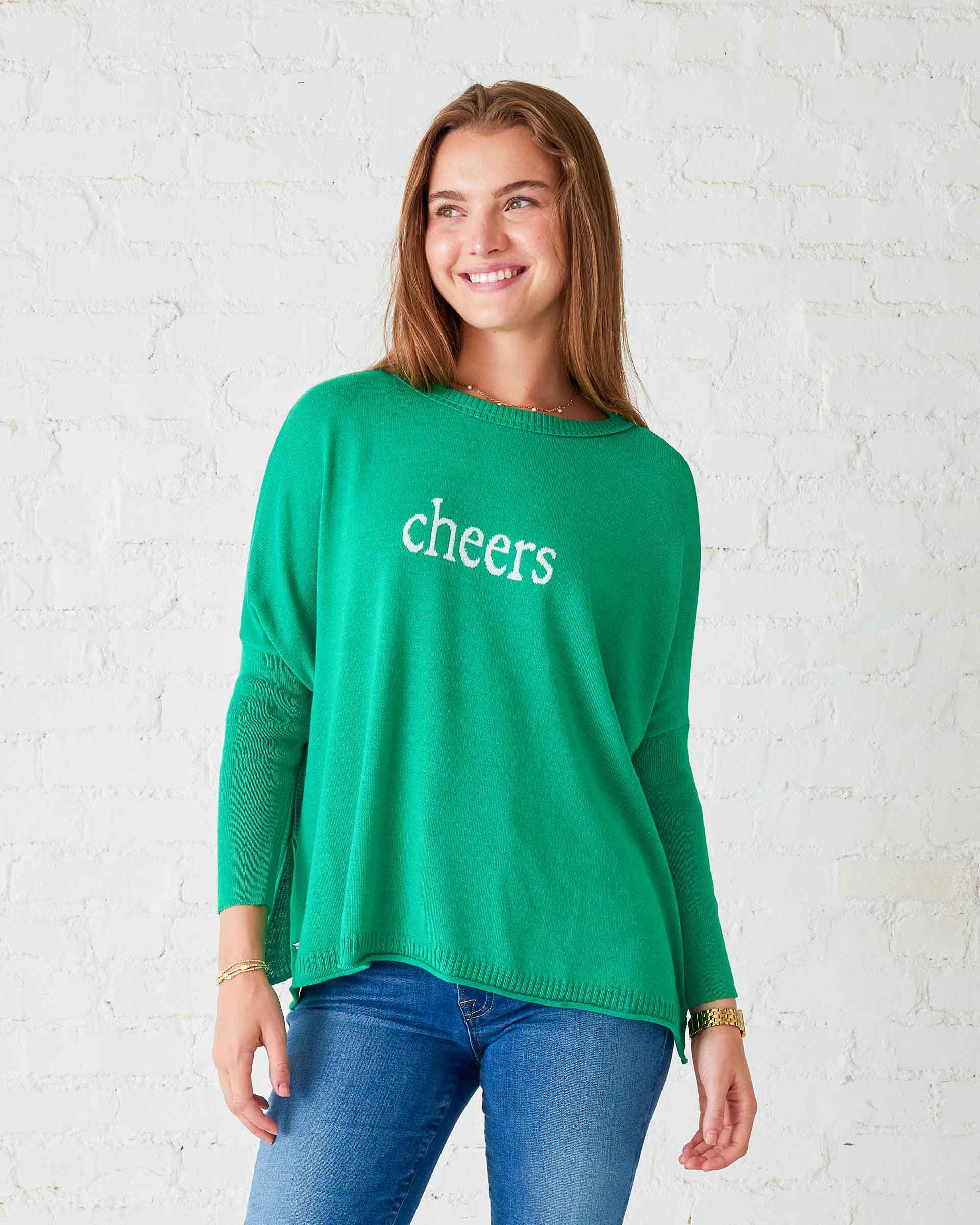 A woman in a versatile green MERSEA - CHEERS CATALINA CREWNECK SWEATER, featuring the word "cheers," stands against a white brick wall, smiling with her hands at her sides. This staple piece from MER SEA pairs perfectly with blue jeans and a gold bracelet.