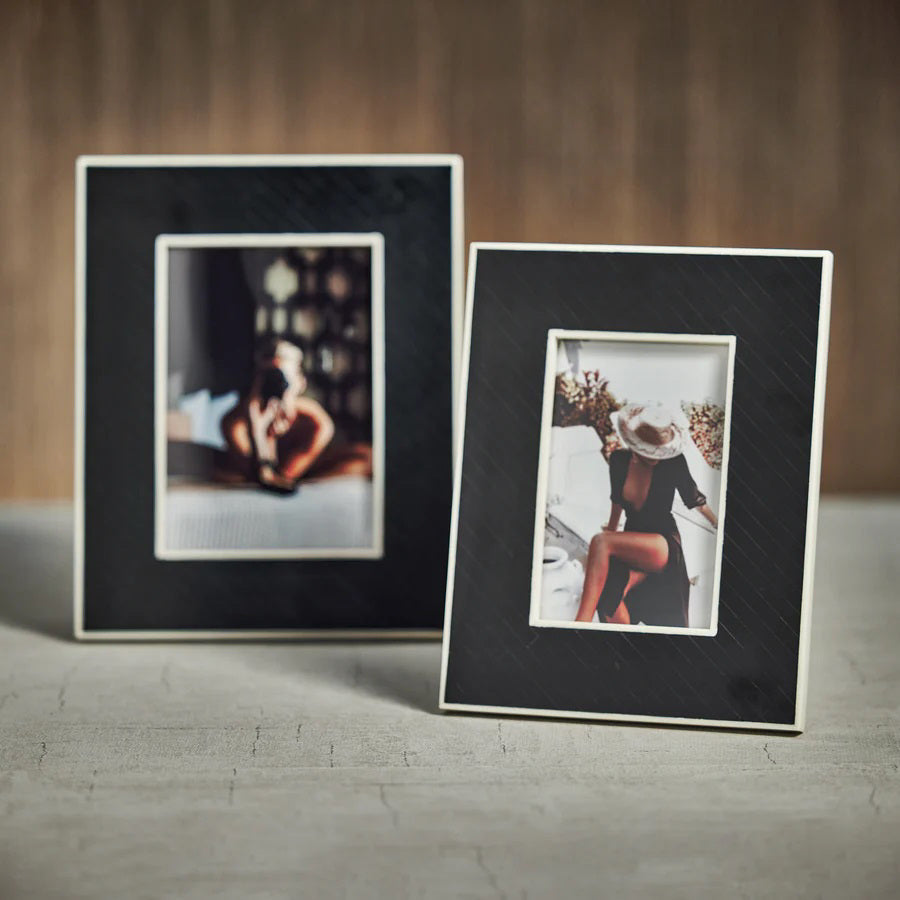 Two framed photographs are displayed on a textured surface. One MDF frame, slightly larger, holds a photo of a person resting on a surface. The smaller frame, from Zodax's BLACK BONE INLAY WITH WHITE BORDER 4X6 collection, contains an image of a person sitting and wearing a hat. Both frames have dark borders with light interiors.