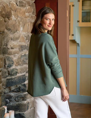 A person with shoulder-length brown hair is wearing the FRANK & EILEEN Effie Long Sleeve Funnel Neck Capelet in rosemary and white pants, smiling by a stone wall in a room with colorful cabinets, exuding the cozy vibe of a winter warmer.