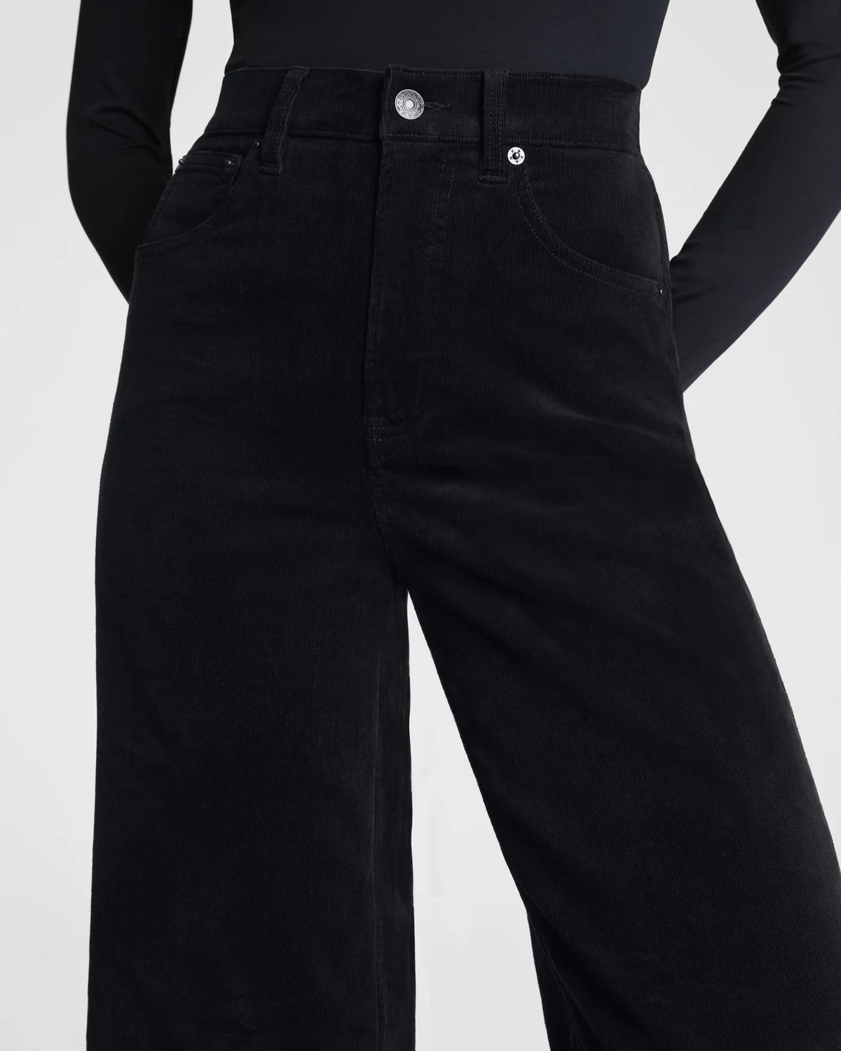 A woman poses against a plain background wearing the SPANX - CORDUROY WIDE LEG JEAN IN BLACK, paired with a fitted black long-sleeve top, black belt, and black heels, her hands in her pockets and expression neutral.