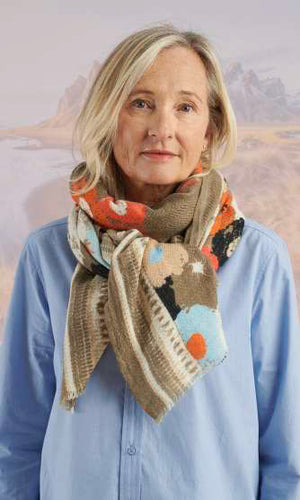 A woman outdoors with a blurred mountain backdrop wears a cream button-up and the STORIATIPIC DORIS Wool Silk Scarf by STORIATIPIC, featuring green, navy, and yellow patterns in a luxe wool-silk blend.
