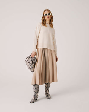 A person with long hair wearing sunglasses poses against a plain background. They wear a cream-colored sweater, a SFIZIO pleated mix media skirt in beige faux leather, and matching knee-high snakeskin boots. They hold a snakeskin-patterned handbag.