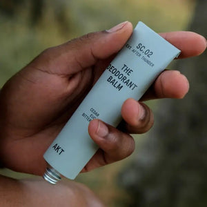 A tube and box of AKT LONDON AKT DEODORANT BALM AFTER THUNDER, featuring cedar, eucalyptus, and orange, provide all-day protection. The minimalist packaging highlights its aluminum-free formula on a beige background.