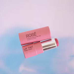 A tube of pink lip tint with the cap partially removed, revealing the tinted balm inside. The tube reads "PINK HOUSE ORGANICS - LIP TINT ROSE" and "LIP TINT BAUME À LÈVRES" against a soft pink and blue background, promising a super hydrating formula in a delicate Rosé shade.