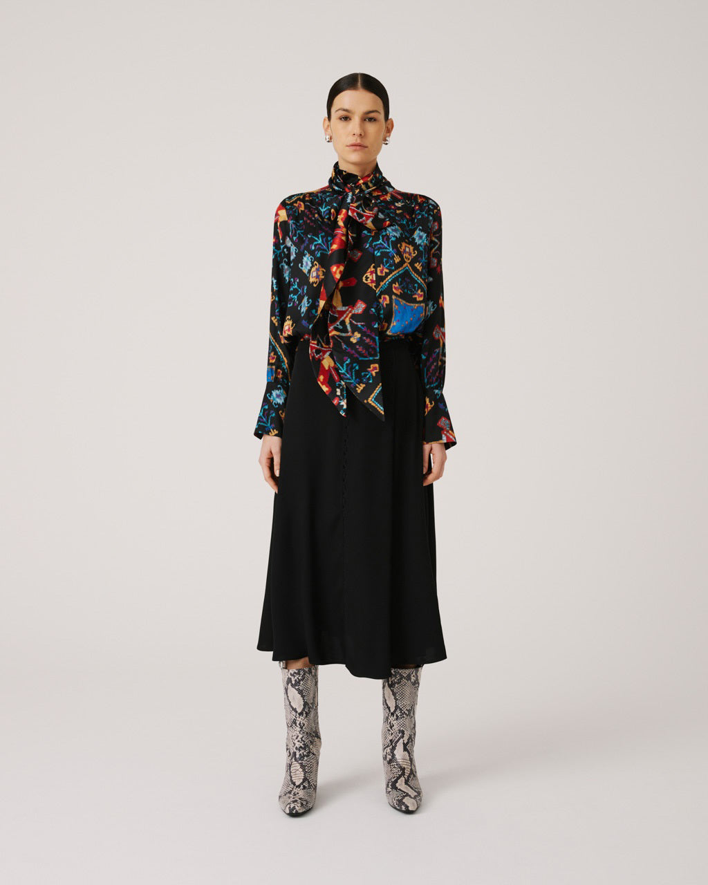 A person with dark hair is standing against a plain background. They are wearing a long-sleeve blouse with a colorful, intricate pattern paired with an elegant SFIZIO - BLACK SKIRT WITH LACE INLAY by SFIZIO. Their outfit, finished with knee-high snakeskin boots made in Italy, looks chic as they look directly at the camera.