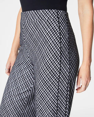 A person is wearing SPANX's Ponte Jacquard Wide Leg Pant, designed with a high-waisted fit and showcasing a black and white grid pattern crafted from premium fabric. They are paired with a black top, standing with their right arm by their side. The focus is on the stylish pants by SPANX.