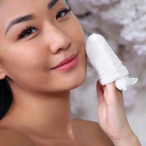 Close-up of a person smiling while gently exfoliating their face with a FACIAL MICRO SCRUBBER from DAILY CONCEPTS, made from white microfiber. The background is blurred, featuring light-colored floral decorations. The person looks relaxed and is displaying smooth, healthy-looking skin after using the light daily facial scrub.