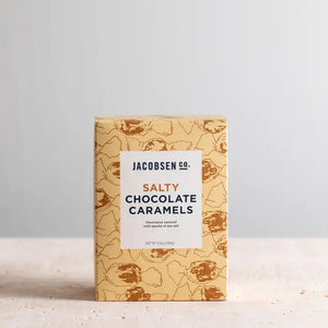 A box of Jacobsen Salt Co. Chocolate Caramels with Sea Salt is shown against a neutral background. The packaging is light blue with a white label featuring the product name in bold letters. The box, highlighting the rich chocolate and sea salt flavors, is decorated with faint illustrations of shells.