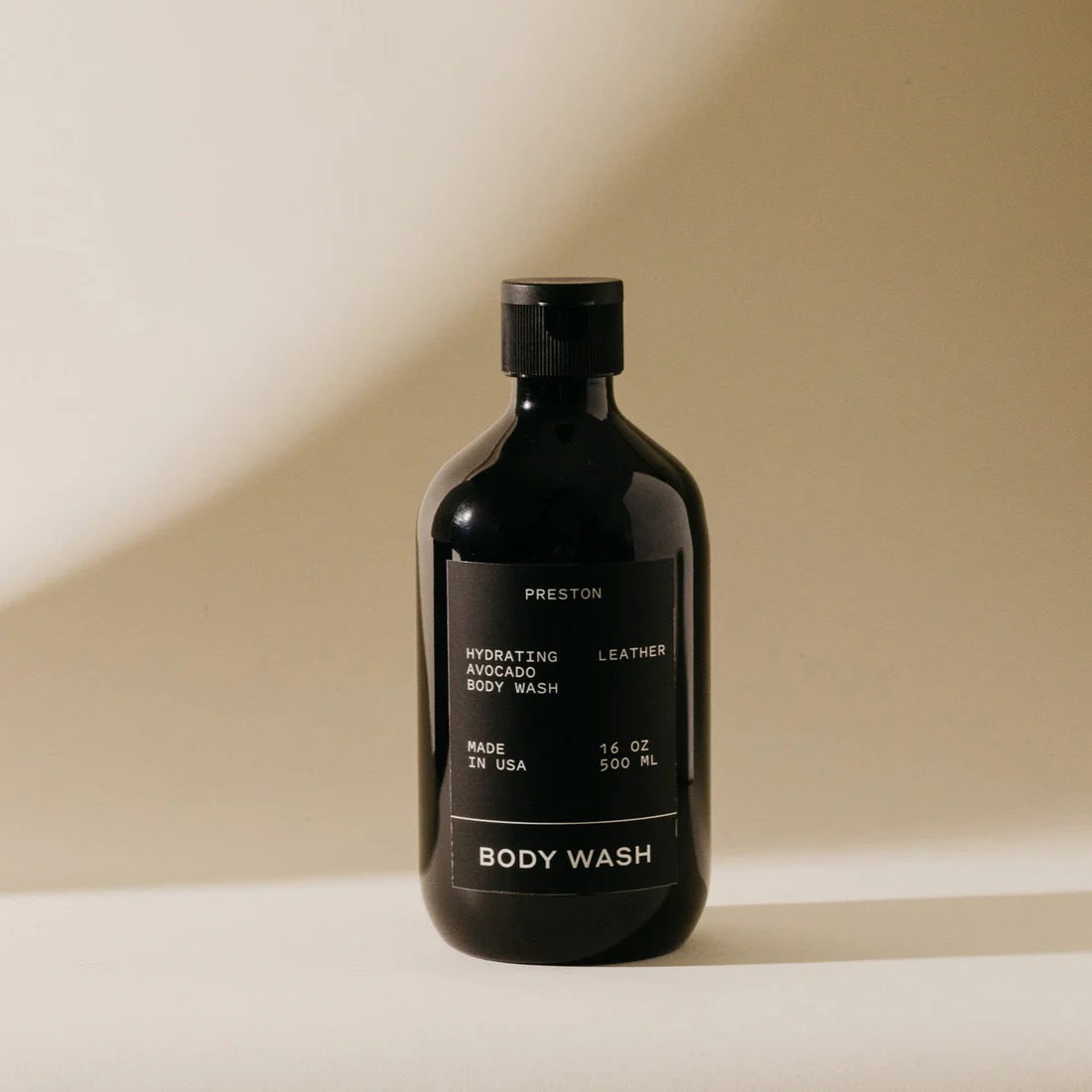 A black bottle of PRESTON - BODY WASH labeled &quot;Hydrating Avocado Body Wash Leather&quot; is centered against a beige background. Made in the USA with plant-based ingredients, this 16 oz (500 ml) moisturizing body wash from the brand PRESTON ensures hydrated skin without harsh chemicals.