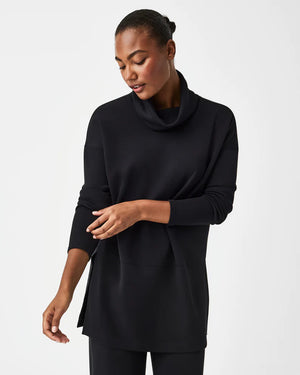 Dressed in the luxurious comfort of a SPANX AireEssentials Turtleneck Tunic, characterized by its black hue and lightweight fabric, a person stands with their back to the camera. Their elegantly tied up hair complements the matching black pants against a plain white backdrop.