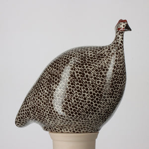 Introducing the LES CERAMIQUES DE LUSSAN - Large Ceramic French Guinea in Black Spotted White by LES CERAMIQUES DE LUSSAN. This exquisite handmade ceramic sculpture features a bird with a speckled pattern, characterized by its round body, small head, and subtle red accents. Reminiscent of the French brood style, it sits gracefully on a smooth beige pedestal against a plain background.