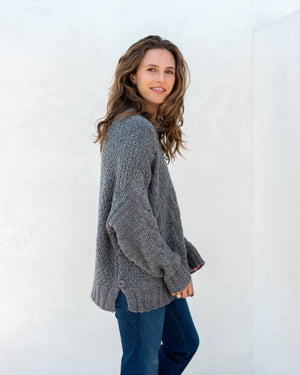 The image displays a detailed view of a person dressed in the MERSEA - TOPANGA CABLE SWEATER by MER SEA, showcasing its chunky knit cream-colored texture with large buttons on the sides, paired with blue jeans. The emphasis is on the sweater's intricate cable design and fine details, with the person's hand partially visible at their side.