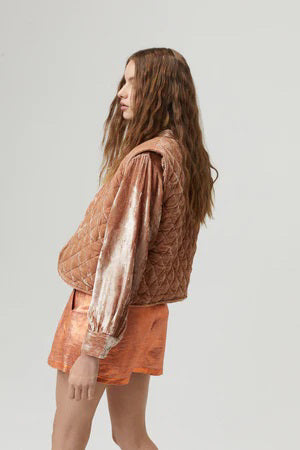 A woman is adorned in a light brown LANHTROPY SUMAC VELVET VEST over a shiny beige and brown blouse, paired with metallic orange shorts. She has long, wavy brown hair and is wearing layered necklaces, standing against a plain light gray background. This layered outfit beautifully harmonizes textures.