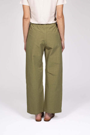 Against a plain background, a person wears THE LITTLE PROJECT's AMBER PANT in olive-green, made from Japanese cotton with a drawstring waist. They're styled with a white short-sleeve top and brown woven flat sandals. These versatile pants are already becoming a best seller.