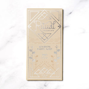 The image shows a light brown rectangular package of Ritual Chocolate labeled "RITUAL CHOCOLATE - BOURBON BARREL AGED CHOCOLATE BAR 75%." This limited edition chocolate, crafted from aged cacao nibs, features geometric designs in silver and white and contains 2.12 oz (60g) of chocolate. The background is a marble surface.