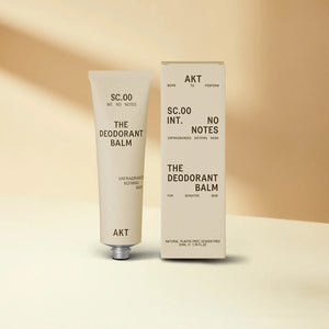 A beige tube of AKT LONDON’s AKT DEODORANT BALM FRAGRANCE FREE for sensitive skin stands upright beside its matching box on a neutral background; both display minimalist black text and design.