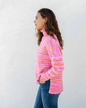 A person wearing the MERSEA - GOOD VIBRATIONS STRIPE TURTLENECK from MER SEA, a cozy sweater made of nylon, polyester, and acrylic in shades of white, gray, and beige, leans against a countertop. They have curly hair and a relaxed posture. The background includes a white vase and several stacked items.