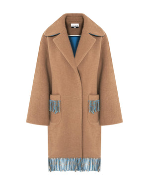 A person is wearing the SFIZIO - TWO TONED COAT WITH FRINGE by SFIZIO, featuring blue and brown fringe patch pockets and hem. They are also donning blue gloves and blue pants. The image is cropped to show from the shoulders down against a plain, off-white background.