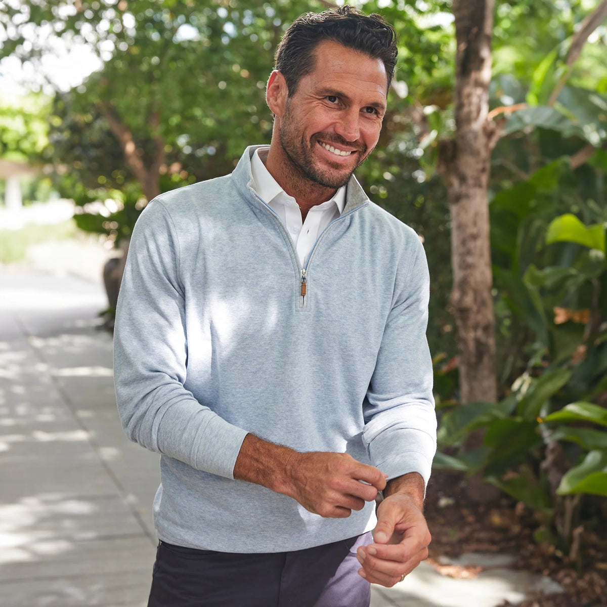 A man smiles outdoors on a sunny day, wearing the COLLARS AND CO Cloud Texture Quarter Zip Pullover in Light Blue over a white polo shirt, with greenery and a shaded path behind him.