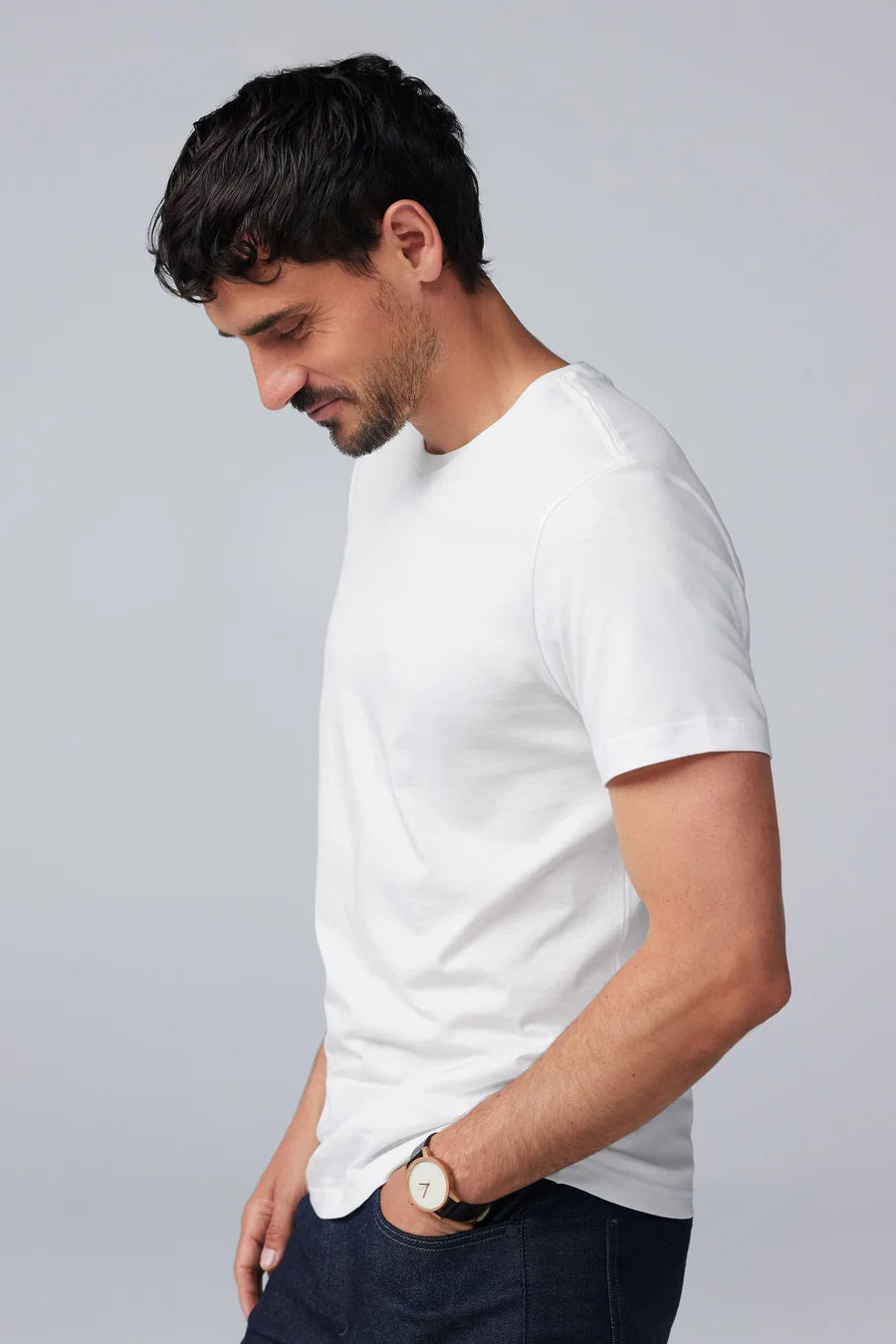 A man with short dark hair and a beard wears a slim fit MACK WELDON SILVER CREW NECK T-SHIRT in white, paired with dark jeans and a wristwatch. He stands before a light gray background, gazing slightly to the side with a neutral expression.