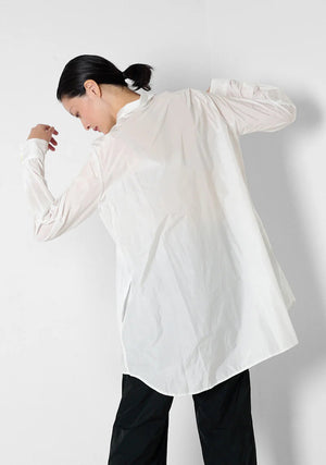 Against a light gray background, a person models the KATHARINA HOVMAN - CLASSIC OVERSIZED SHIRT with mother-of-pearl buttons and black pants. Their hair is tied back as they face away, slightly raising their arms to highlight the shirt's loose fit.