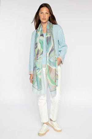 A person with long hair elegantly wears a light blue sweater, white pants, and sleek white shoes. Gracefully draped around their neck is the palm print scarf from KINROSS CASHMERE by KINROSS against a plain white background.