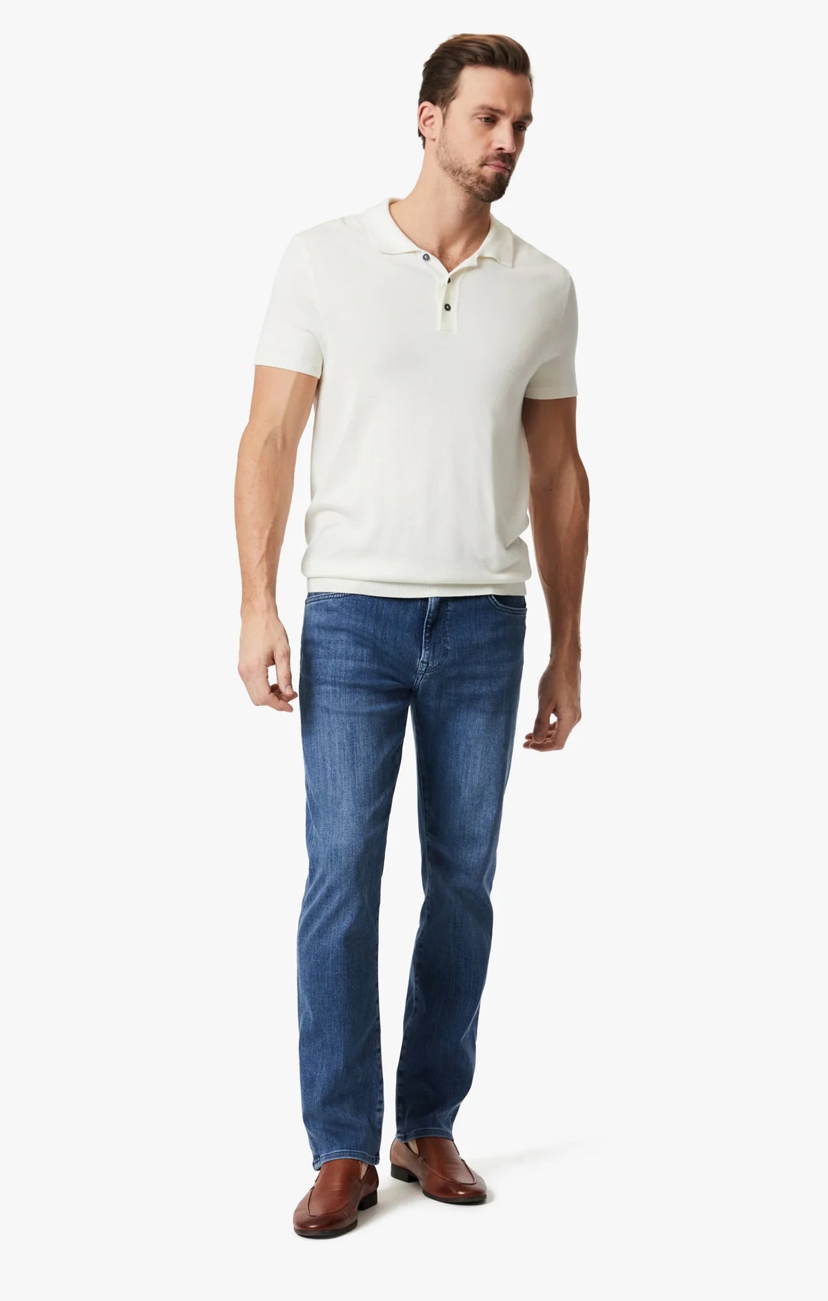 A man wears 34 HERITAGE CHARISMA Relaxed Straight Leg Denim in Mid Urban Wash, a white shirt, and brown leather shoes, standing against a plain white background.