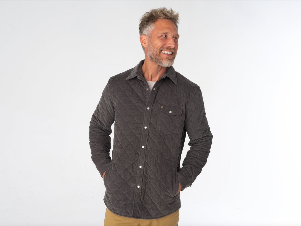 A man with short, curly hair and a beard wears the CRIQUET - CORDUROY QUILTED SHACKET by CRIQUET and blue jeans, standing against a white background while smiling and looking to his right.