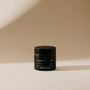 A small black container of PRESTON's cruelty-free beard balm is placed against a neutral, softly-lit backdrop. The label reads "PRESTON - BEARD BALM" along with other details in light/white text. Shadows fall to the right, adding depth to the minimalist design setting that showcases its satin finish.