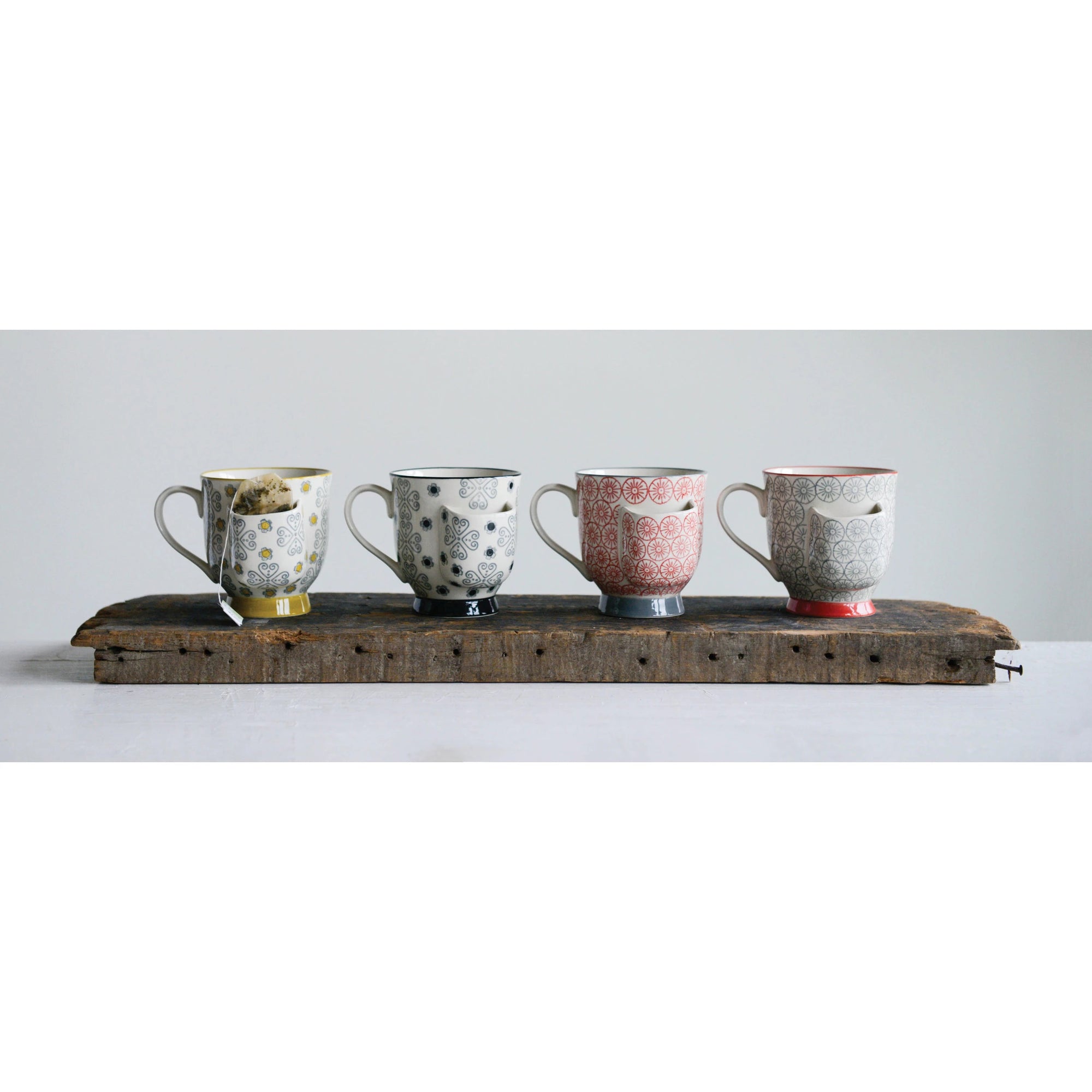 Four intricately patterned stoneware mugs from Creative Coop, each with a unique color, are lined up on a wooden plank against a plain background. Their unique designs create a visually appealing arrangement and, as the "Mug with Tea Bag Holder," they are microwave safe for practicality.