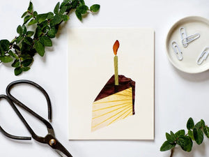 This charming CAKE BIRTHDAY CARD by PETAL PEOPLE PRESS showcases an illustration of a single slice of chocolate frosted cake, adorned with a vertical candle and flickering flame. The simple elegance against a white background evokes the charm of pressed flower art.