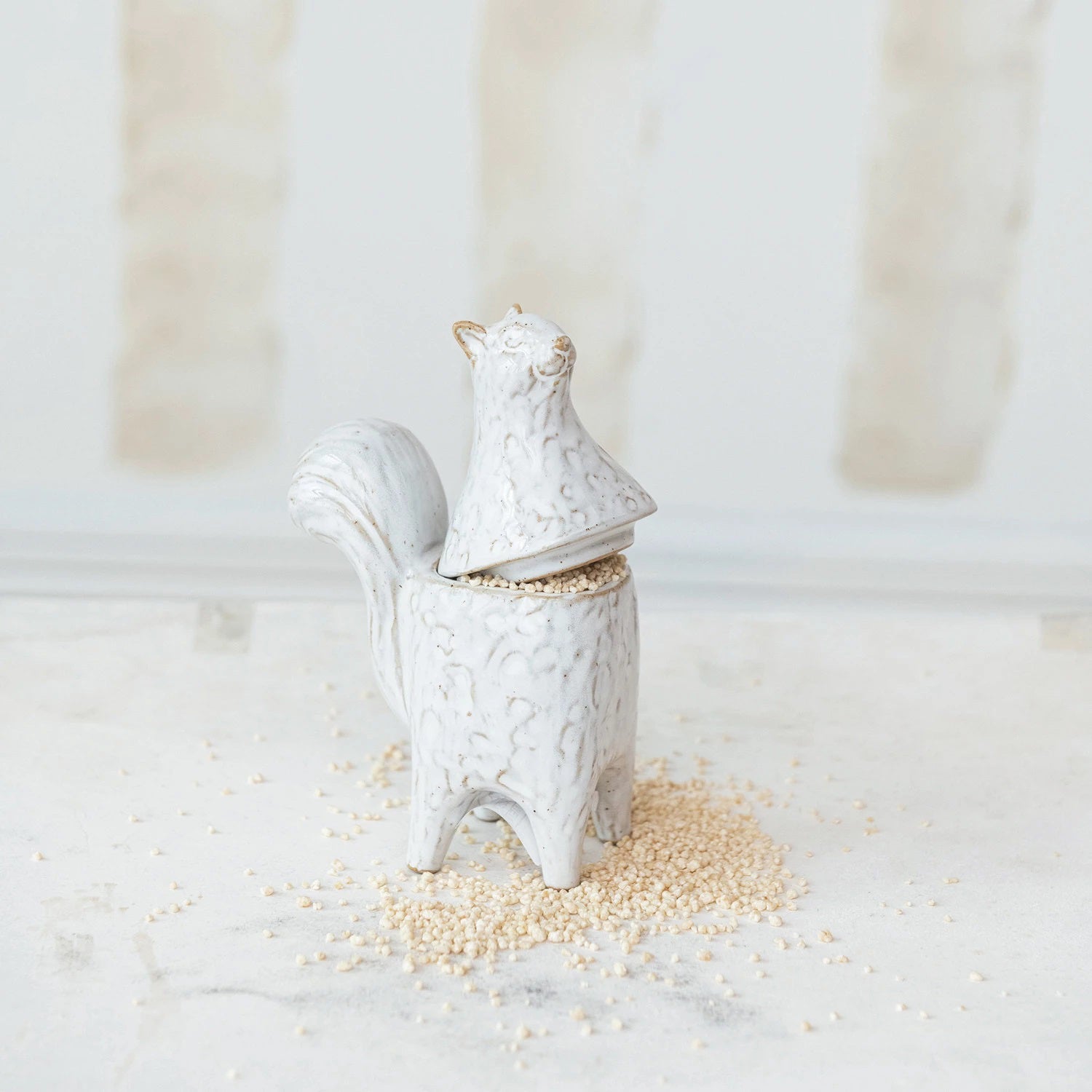 A uniquely-designed STONEWARE SQUIRREL SHAPED JAR from CREATIVE COOP stands on a light surface, with scattered grains or seeds around it. The squirrel's back opens as a lid to reveal the top of the container adorned with reactive glaze. The background is softly blurred in neutral tones.