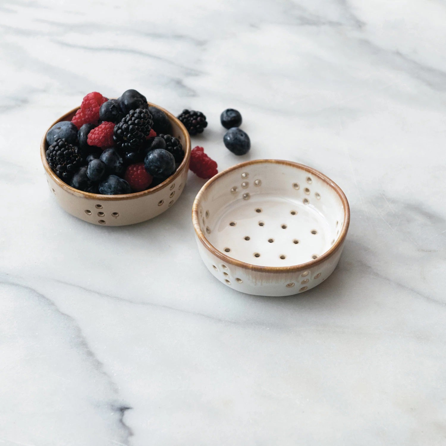 Four Creative Coop STONEWARE BERRY BOWLs, each measuring 4" in diameter, are displayed against a white background. The two bowls on the left feature a white reactive glaze with brown rims, while the two on the right are entirely brown. Each pair includes both a top-down and side view of these ceramic colander bowls with holes.