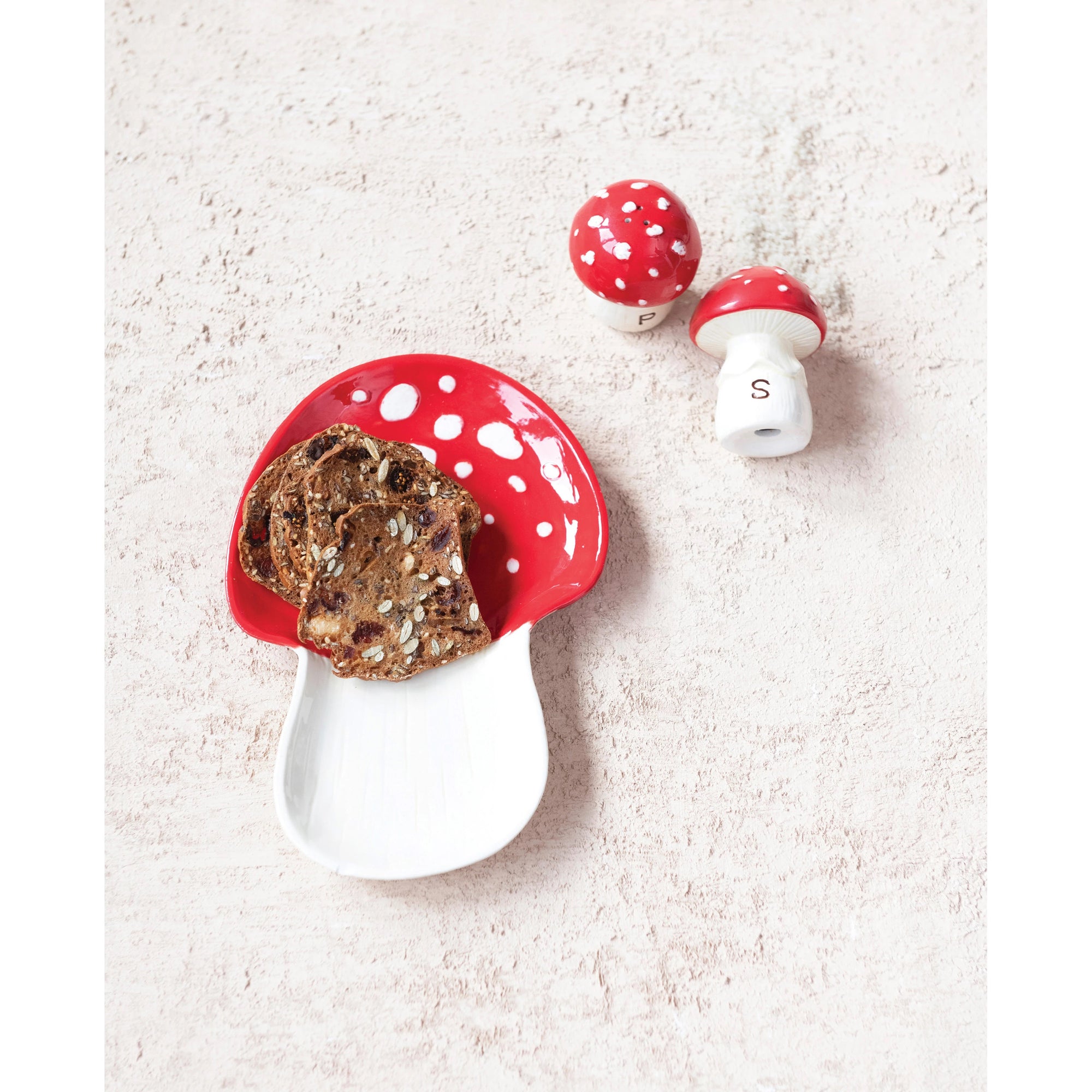 On a textured white surface, there is a serving dish shaped like a mushroom accompanied by crackers. Nearby are two hand-painted ceramic salt and pepper shakers from Creative Coop, each designed in the shape of mushrooms. One shaker features a red cap with white polka dots labeled "P" for pepper, while the other is similarly styled and labeled "S" for salt.